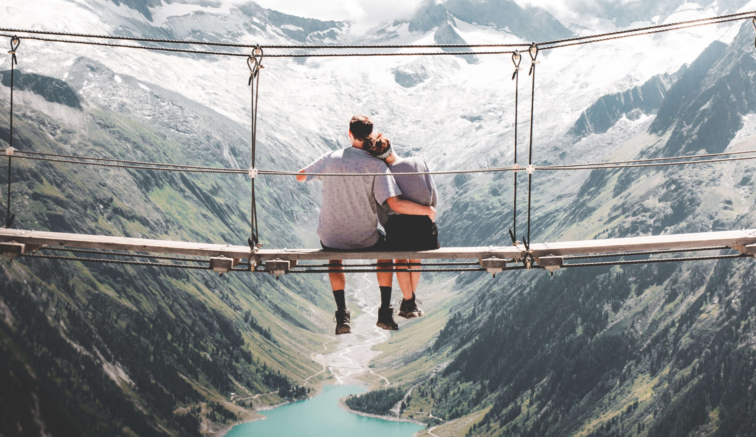 Liebespaar auf einer Brücke Liebespaar, umarmt sich und sitzt auf einer hohen Hängebrücke in den Bergen mit Blick auf einen Gletscher und Bergsee. Es ist Sommer.